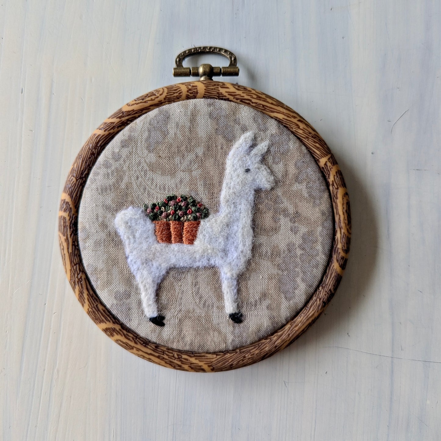 White llama needle felt art with a flower basket on a grey background