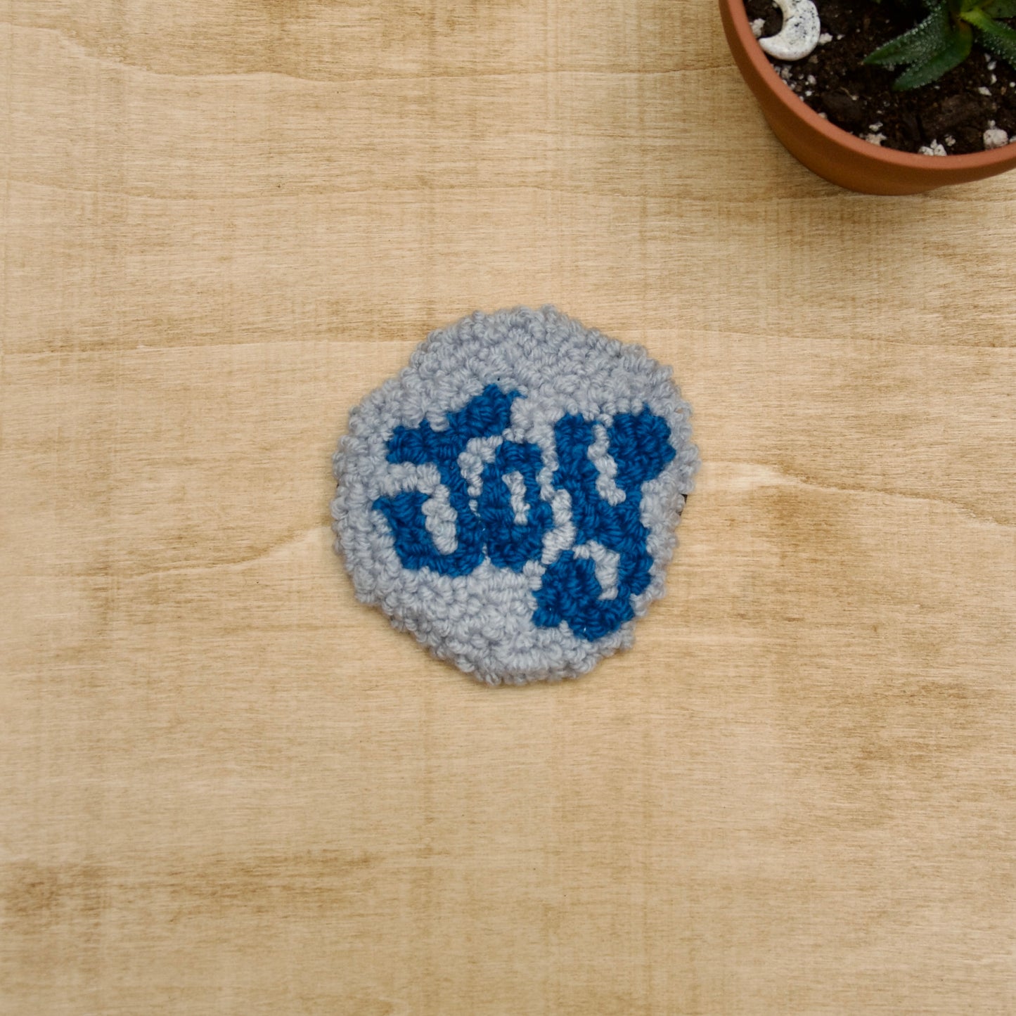 A hand-stitched, circular coaster with the words 'JOY' written on it, placed on a beige surface with a potted plant in the background.