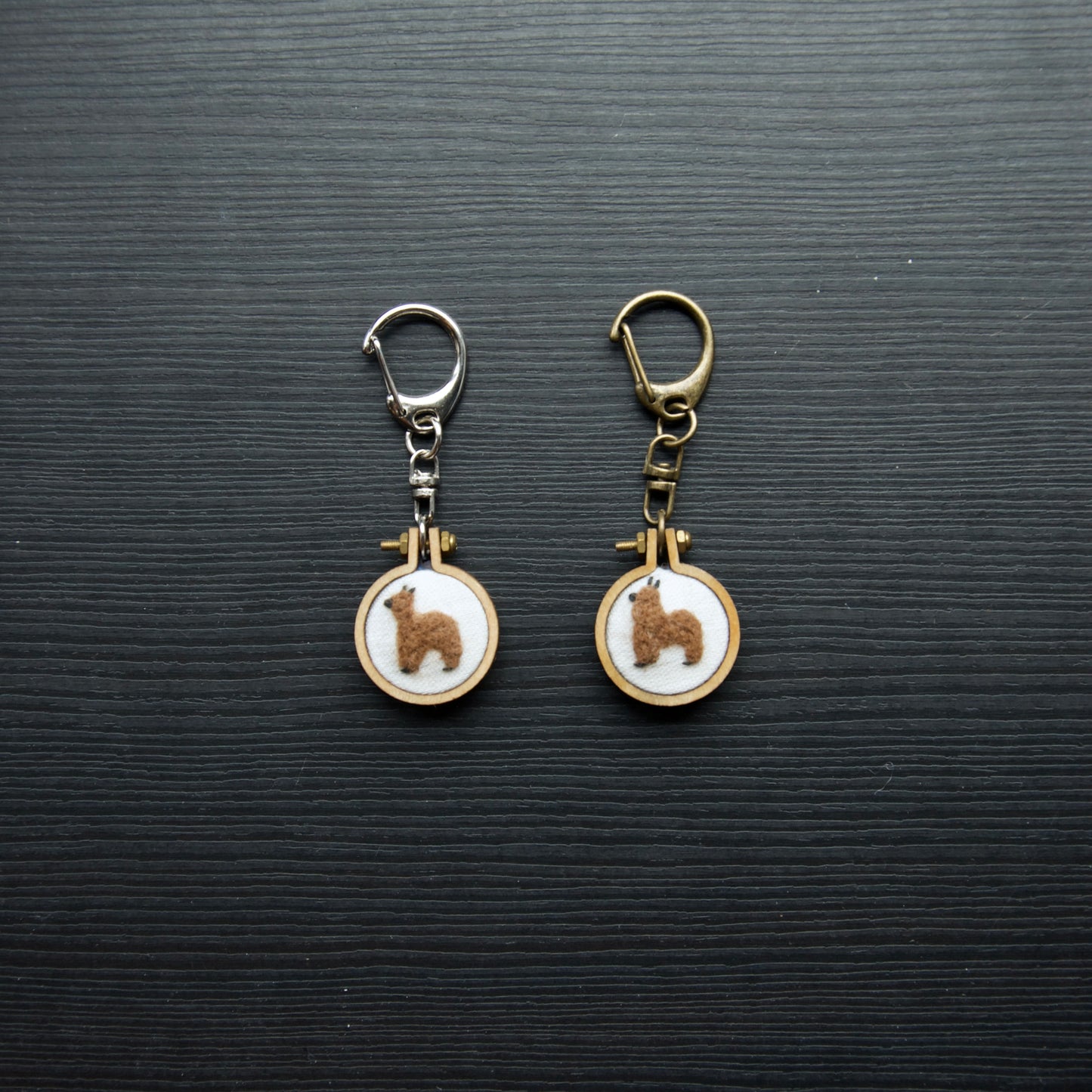 Two needle felt alpacas on small wooden keychains on a dark surface