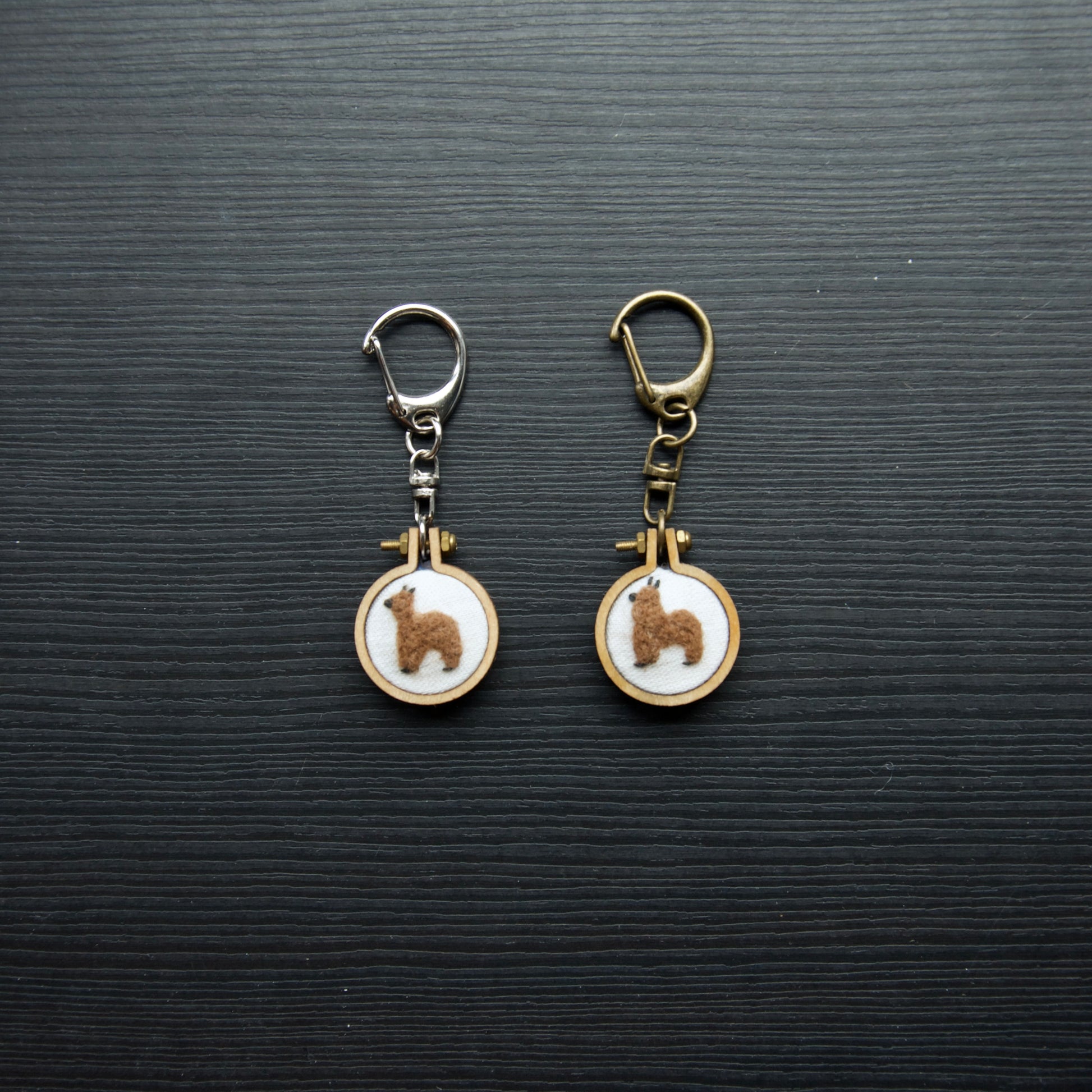 Two needle felt alpacas on small wooden keychains on a dark surface