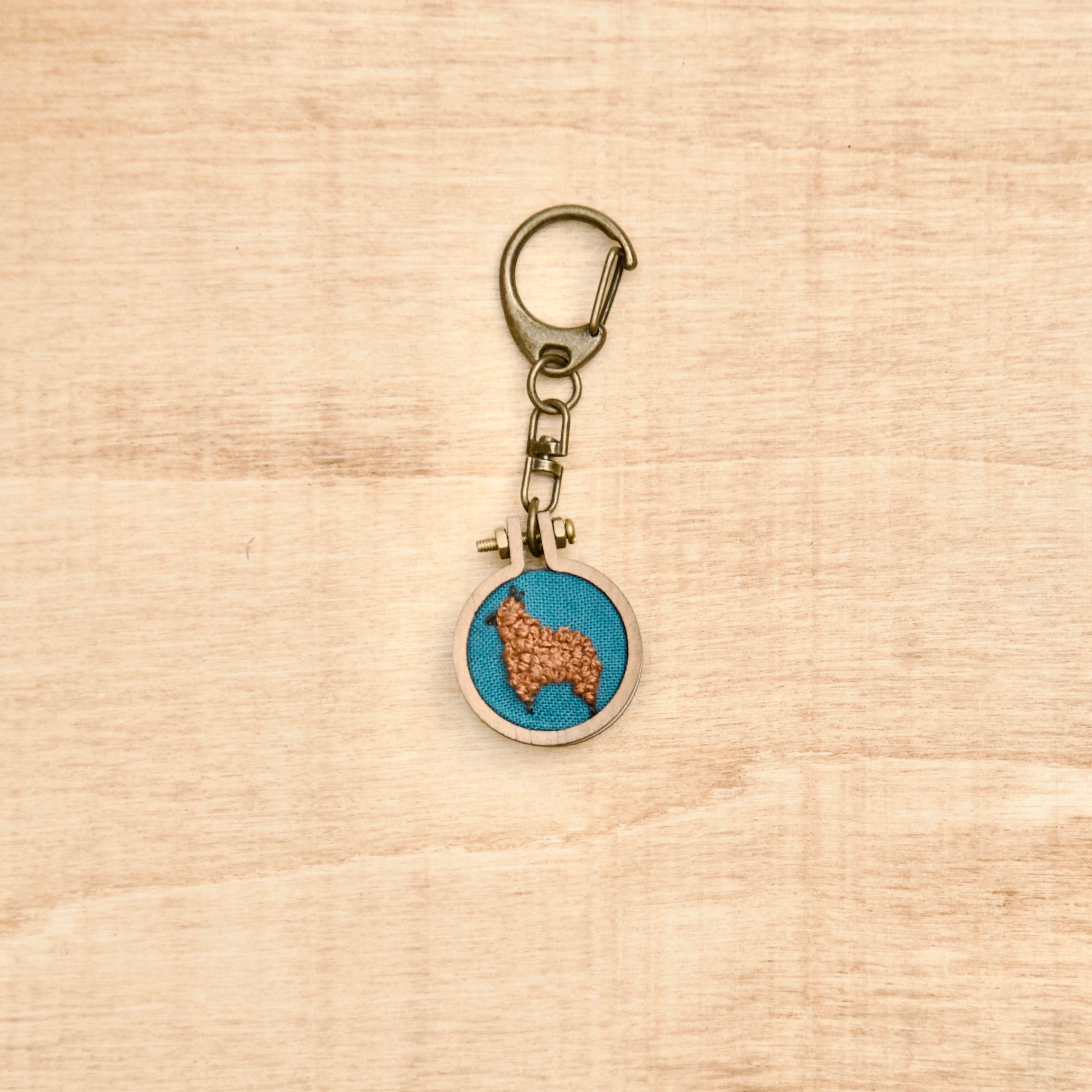 Round keychain with a small embroidered alpaca design on a wood surface with a potted plant nearby