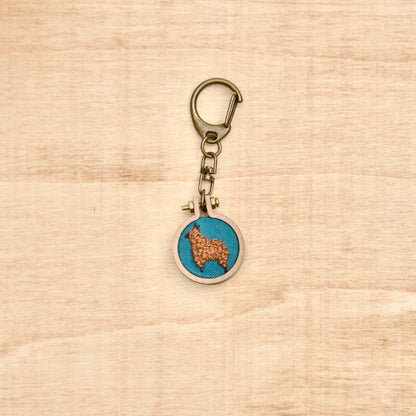 Round keychain with a small embroidered alpaca design on a wood surface with a potted plant nearby