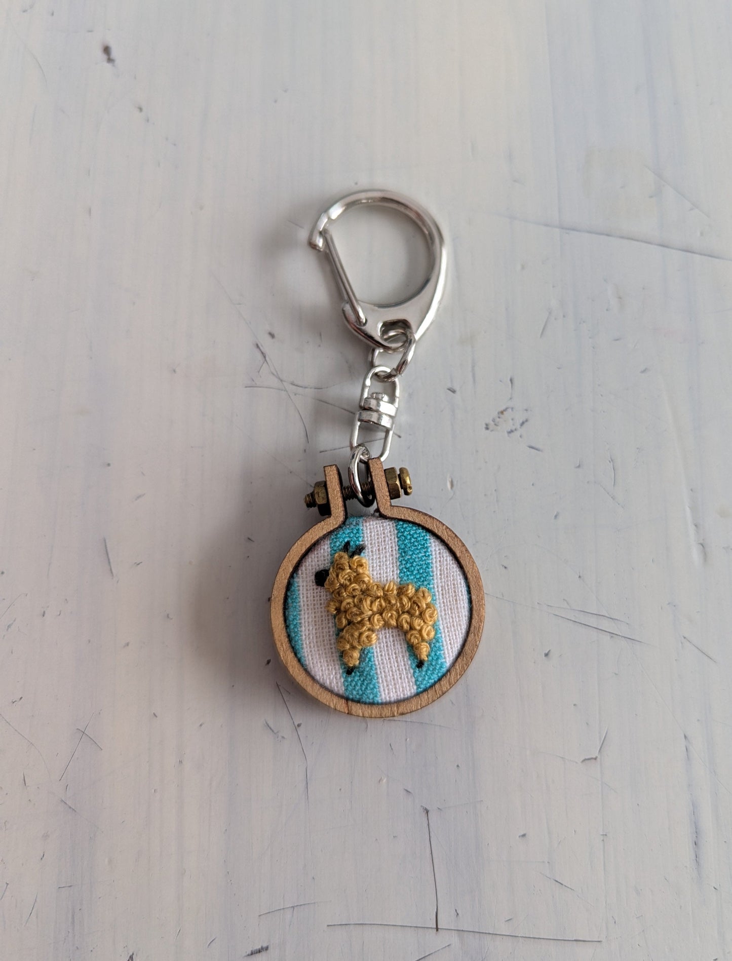 Keychain with embroidered design of an alpaca on a striped background, attached to a metal clasp.