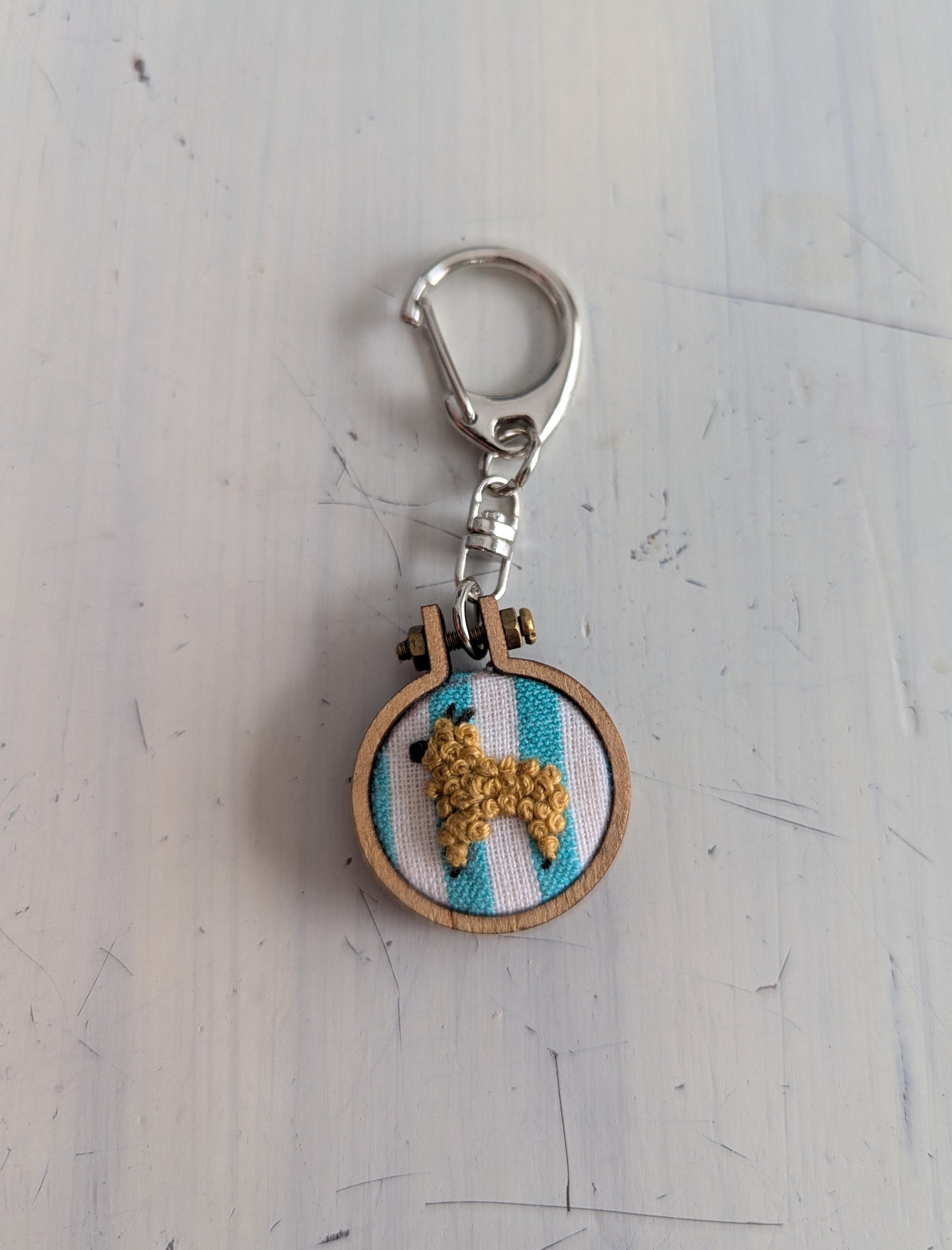 Keychain with embroidered design of an alpaca on a striped background, attached to a metal clasp.
