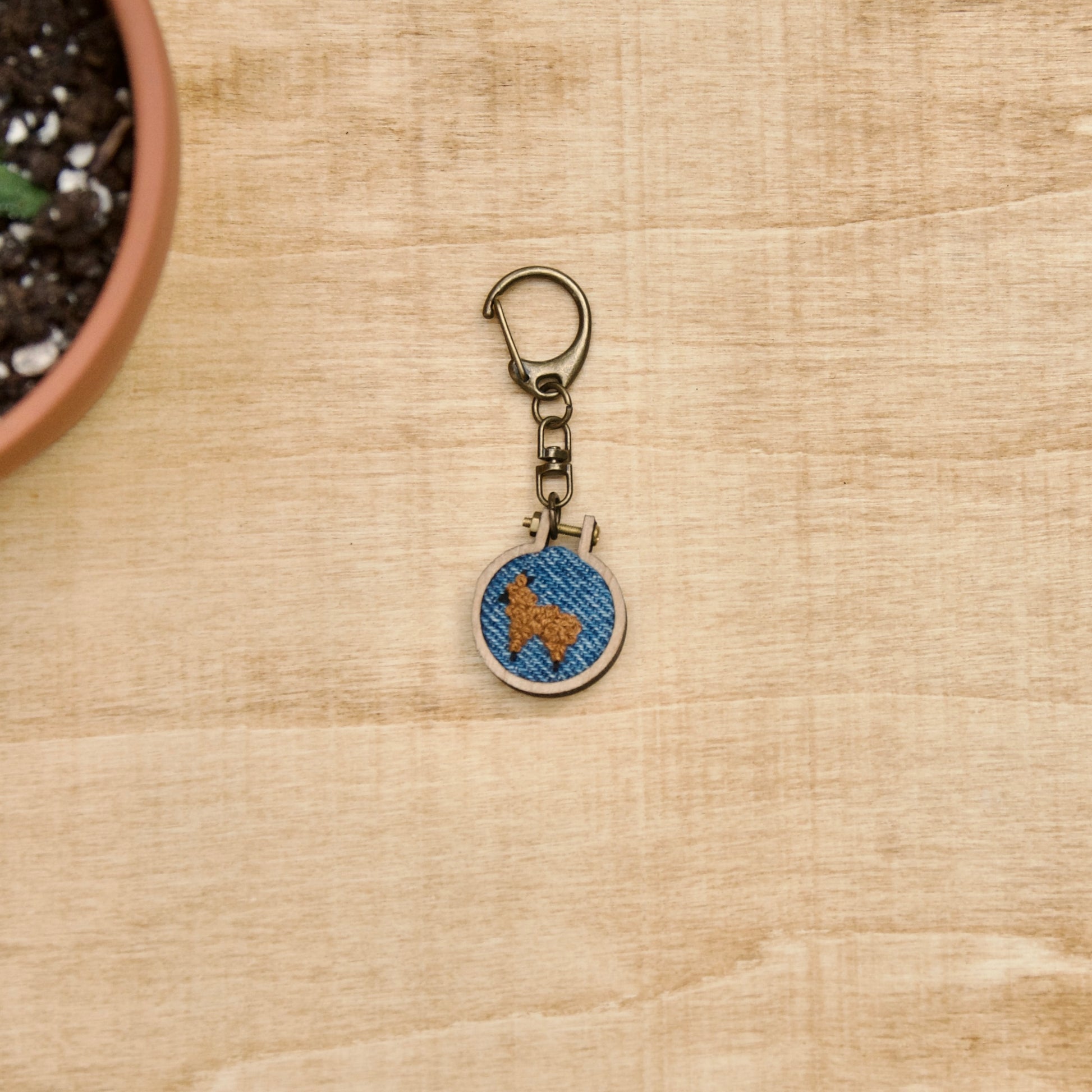 Round keychain with a small embroidered alpaca design on a wood surface with a potted plant nearby