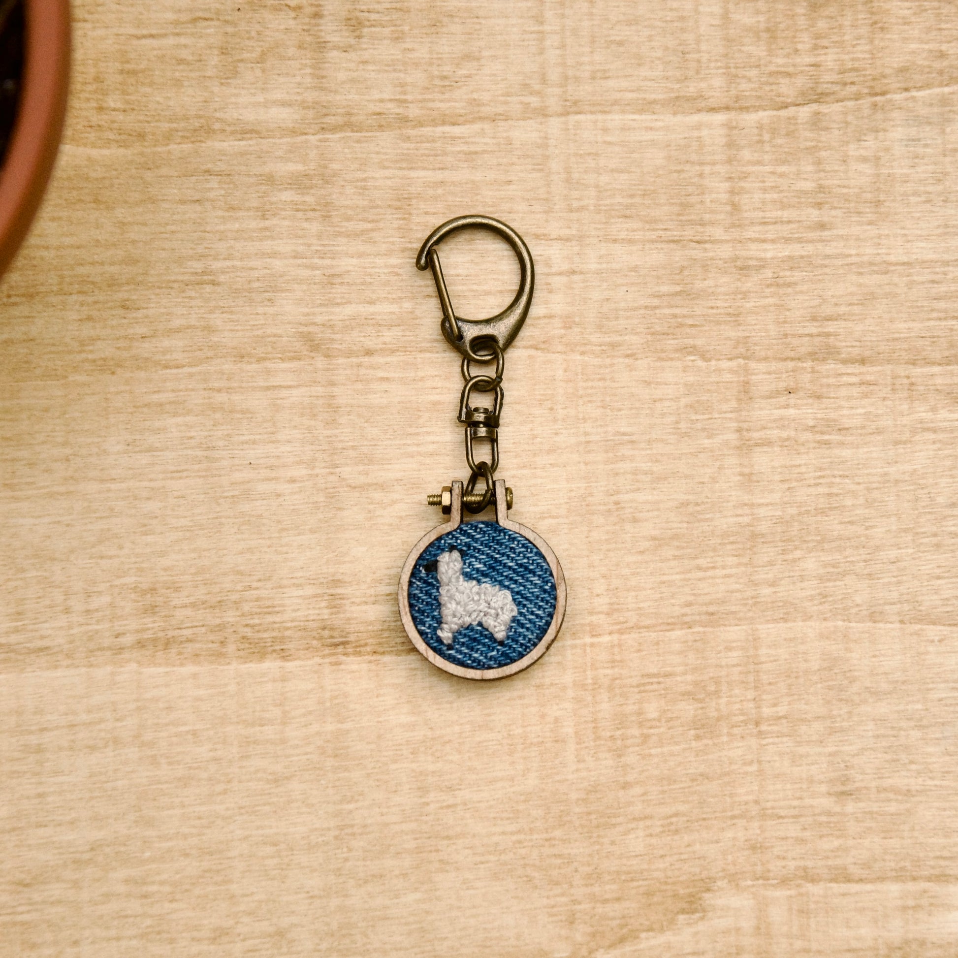 Round keychain with a small embroidered alpaca design on a wood surface with a potted plant nearby