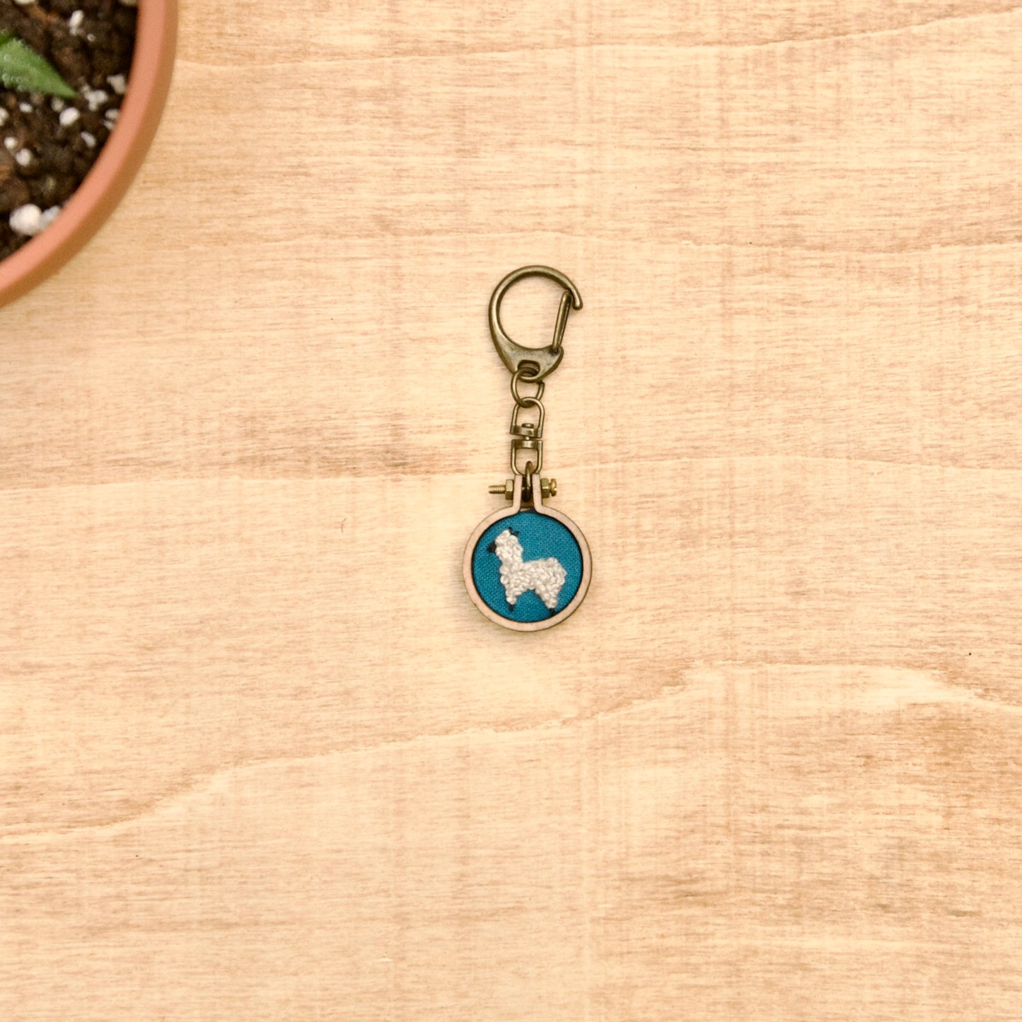 Round keychain with a small embroidered alpaca design on a wood surface with a potted plant nearby