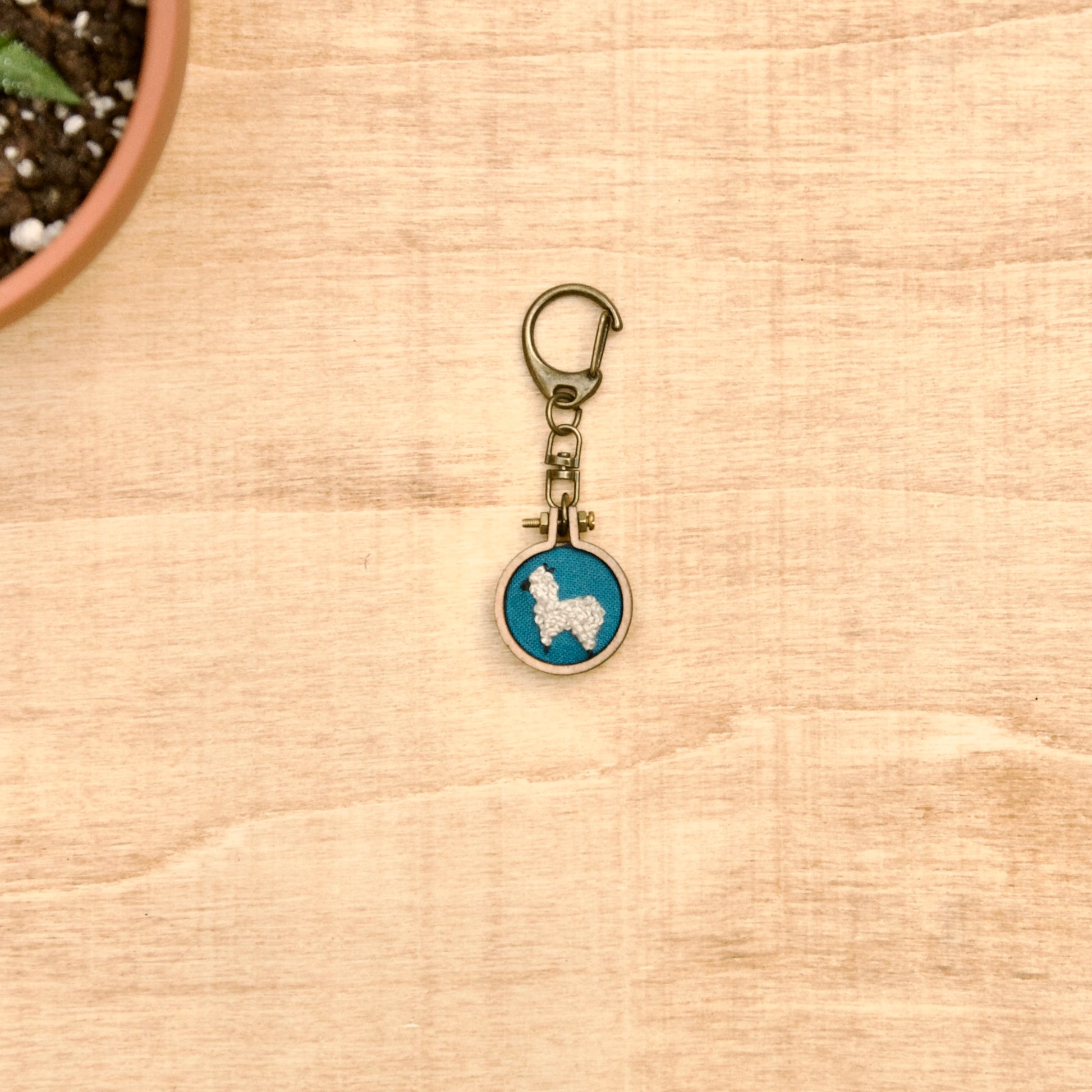 Round keychain with a small embroidered alpaca design on a wood surface with a potted plant nearby