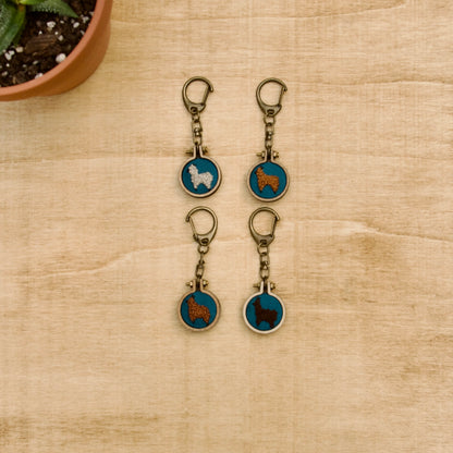Four round keychains with a small embroidered alpaca designs on a wood surface with a potted plant nearby