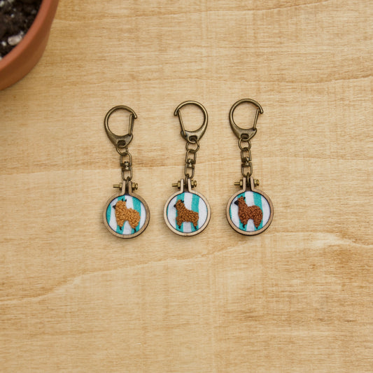 Three embroidered keychains with alpaca designs on a teal stripe background, displayed on a wooden surface.