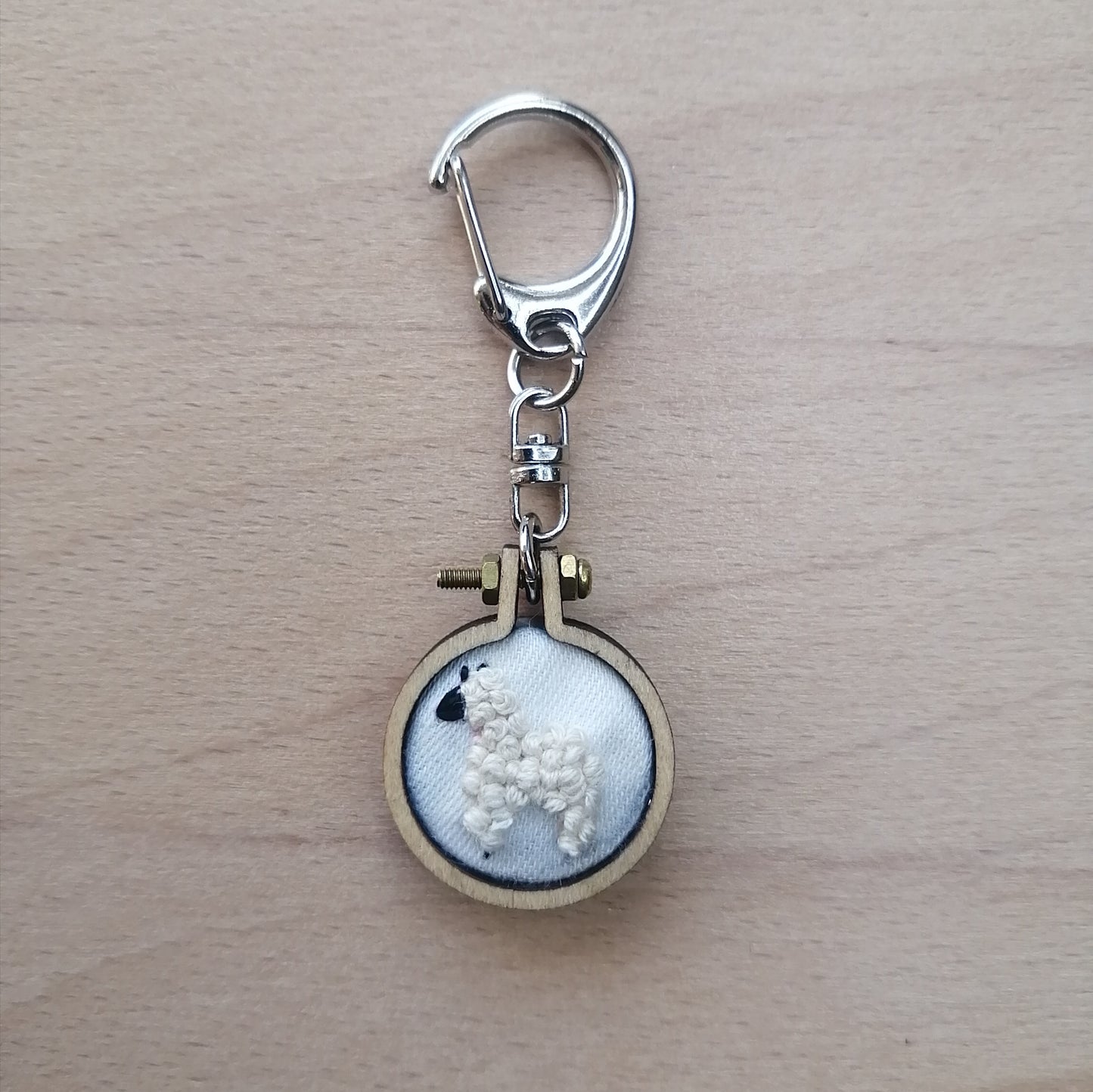 Round keychain with a small embroidered alpaca design on a wood surface with a potted plant nearby