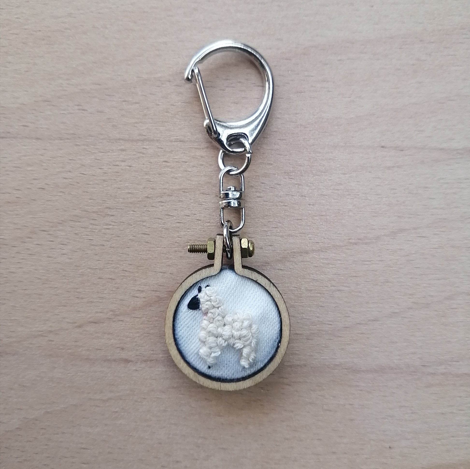 Round keychain with a small embroidered alpaca design on a wood surface with a potted plant nearby