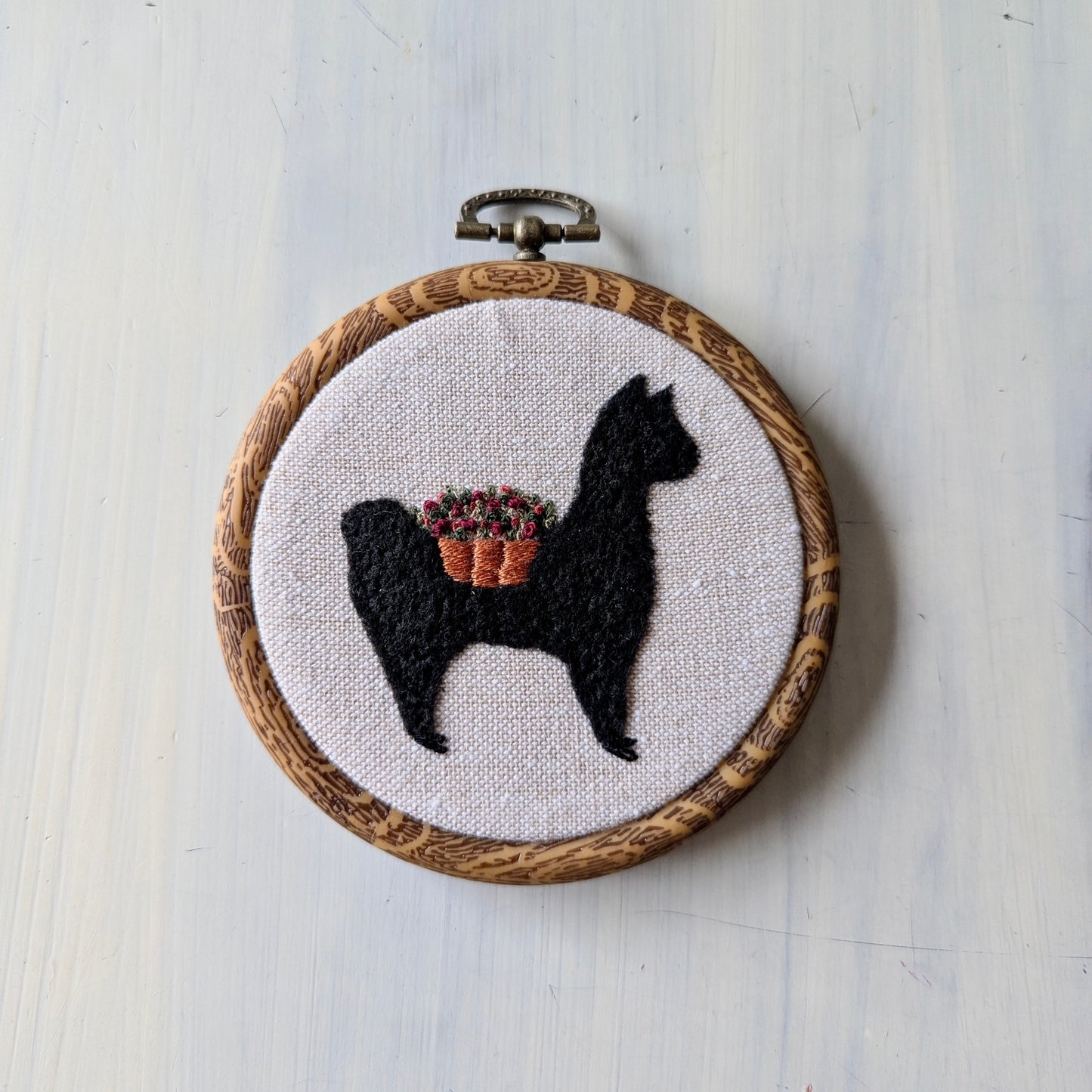 Embroidery hoop with a black alpaca silhouette and flower basket on white fabric, on a light wooden surface.