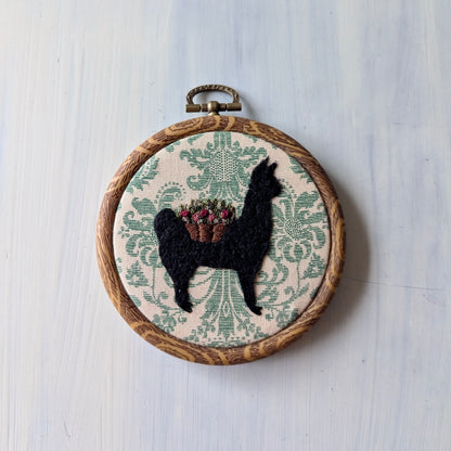 Embroidery hoop with black silhouette of an alpaca wearing a flower basket on a patterned fabric background
