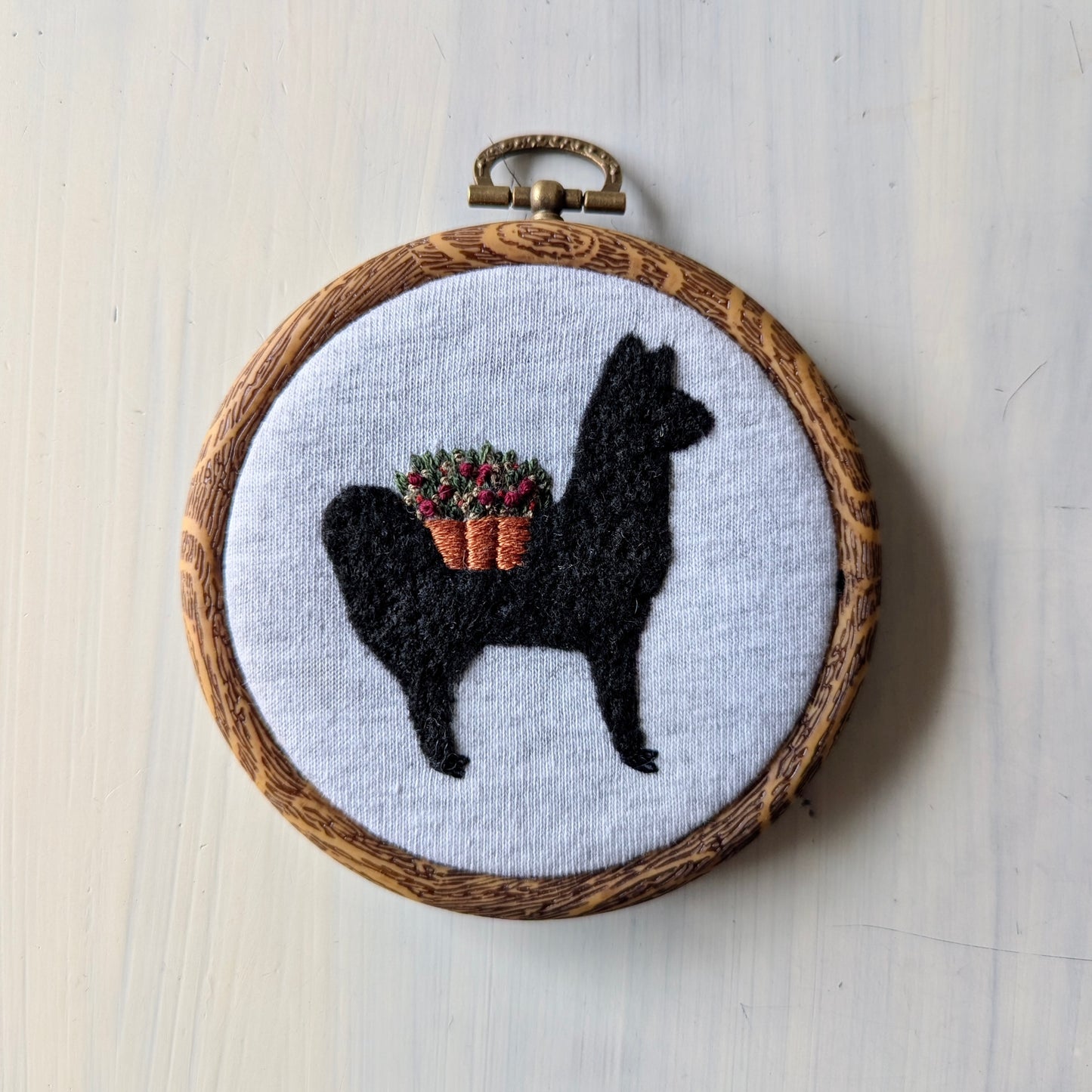 Embroidery hoop with a black alpaca silhouette holding a basket of flowers on a light wooden background