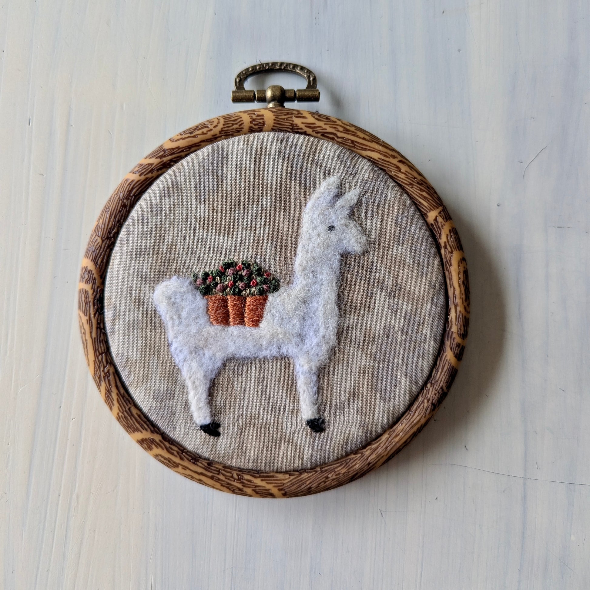 White llama needle felt art with a flower basket on a grey background