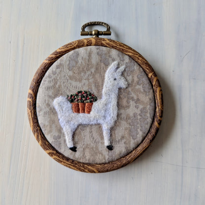 White llama needle felt art with a flower basket on a grey background