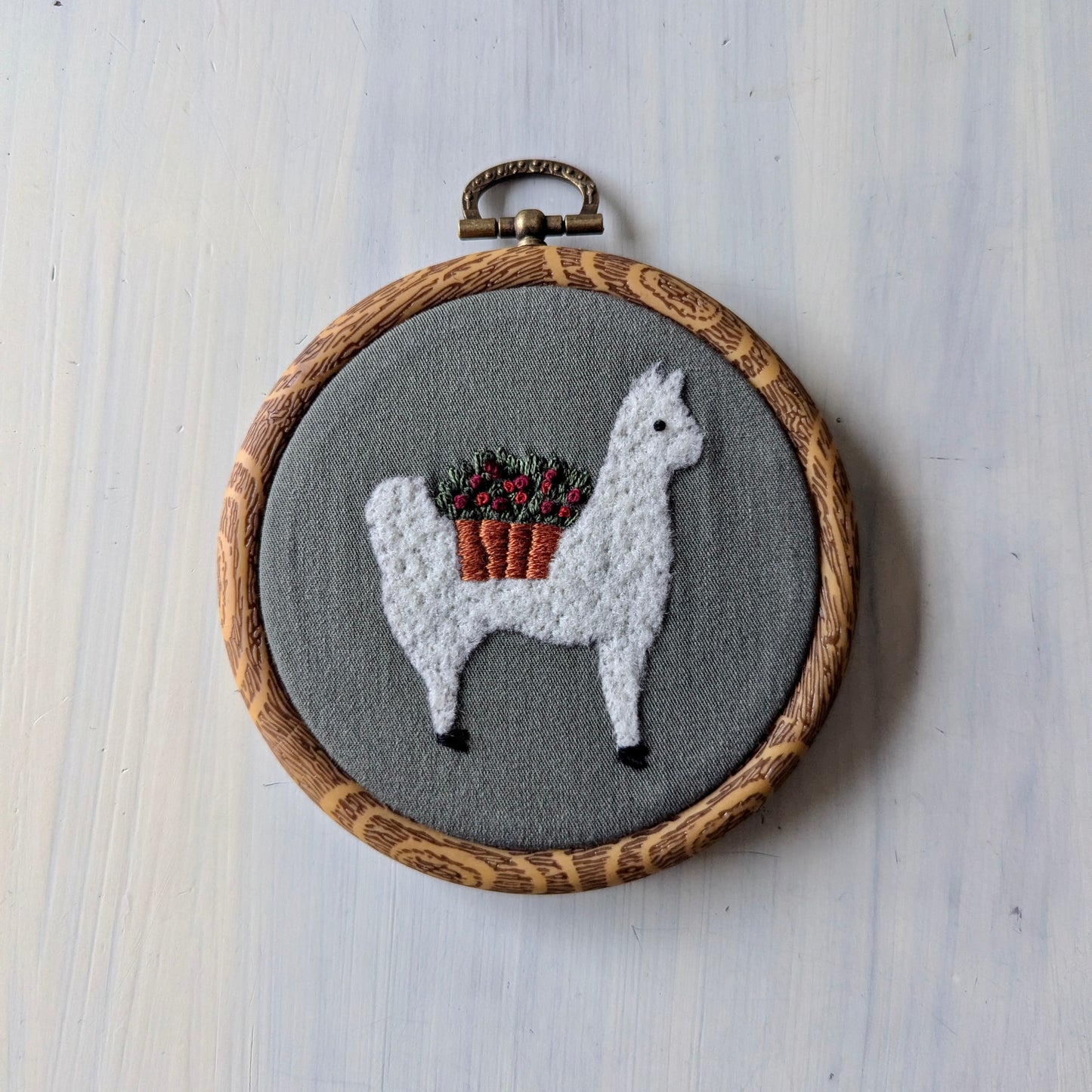Embroidery hoop with a llama wearing a flower basket on olive green fabric, in a wooden embroidery frame
