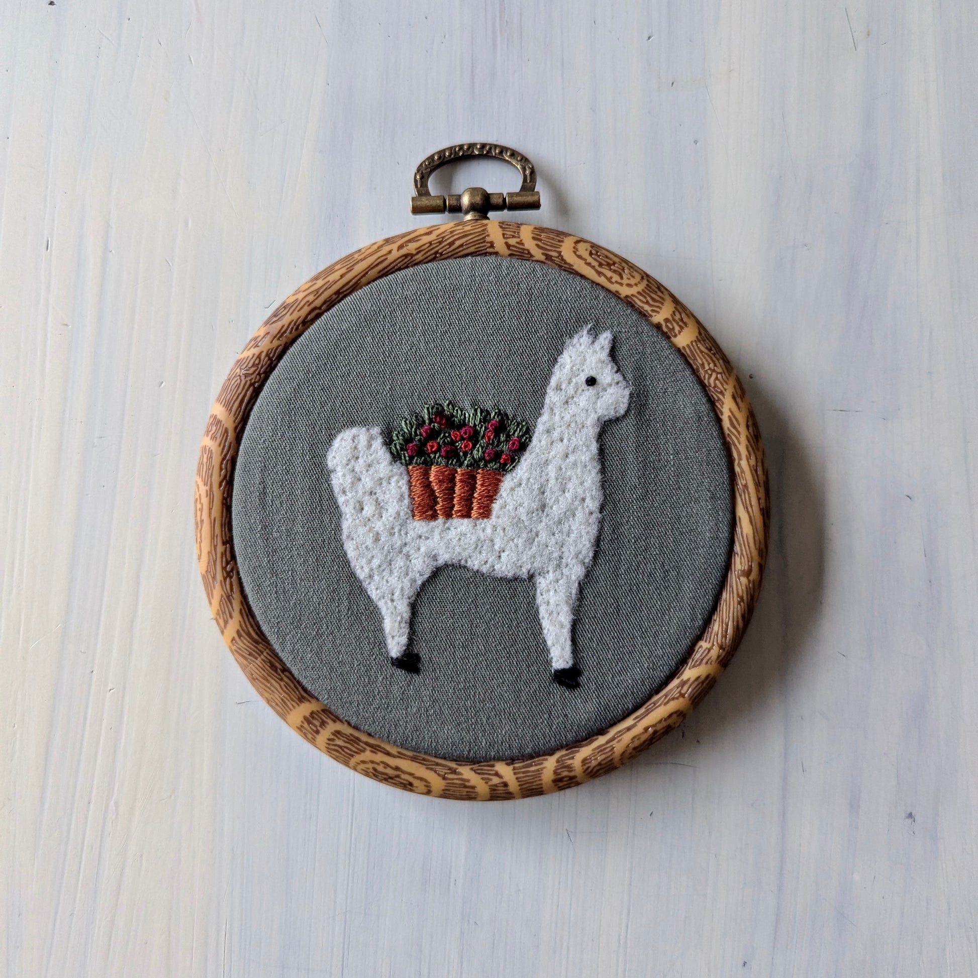 Embroidery hoop with a llama wearing a flower basket on olive green fabric, in a wooden embroidery frame