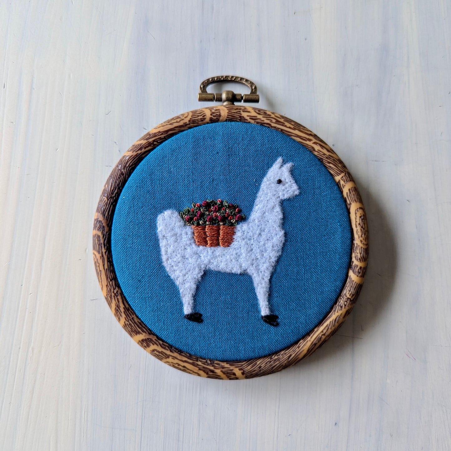 White alpaca wearing a flower basket set over a teal background, in a wooden embroidery hoop