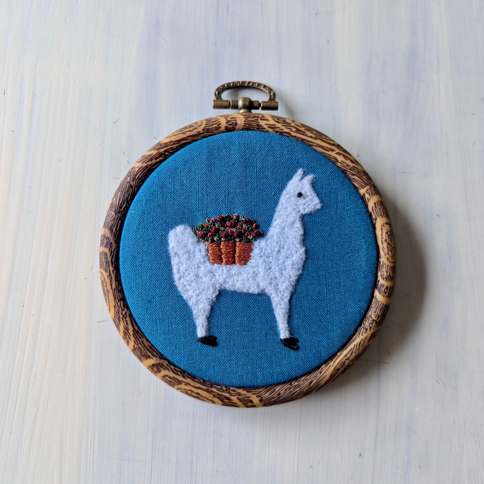 White alpaca wearing a flower basket set over a teal background, in a wooden embroidery hoop