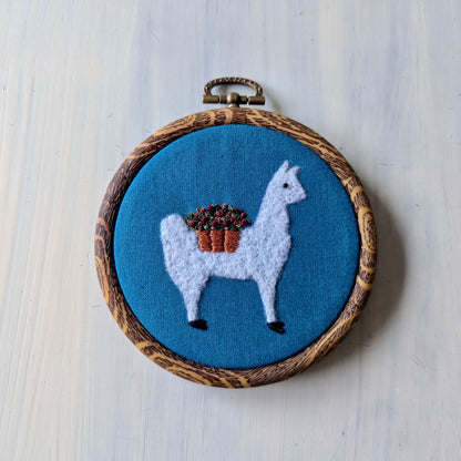 White alpaca wearing a flower basket set over a teal background, in a wooden embroidery hoop