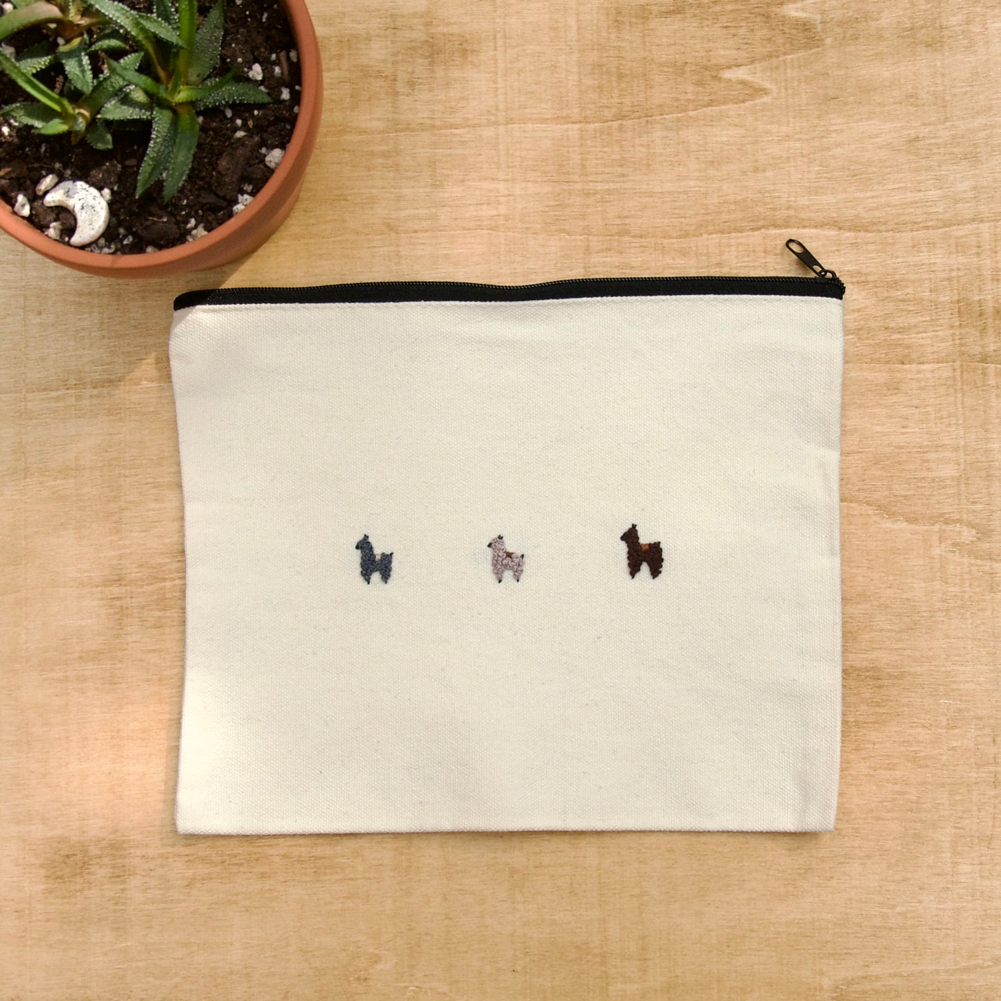 A white zip pouch with embroidered alpacas on the front, displayed on a wooden surface next to a potted plant.