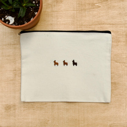 A white zip pouch with 3 brown embroidery alpacas on a wooden surface with a potted plant nearby