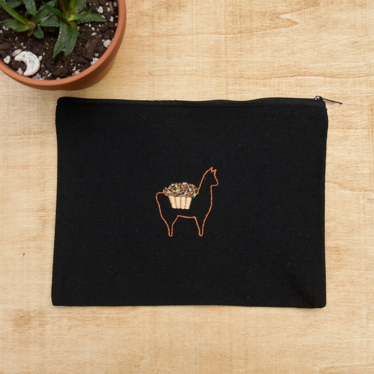 A black zip pouch with an embroidery alpaca with a flower basket on its' back  on a wooden surface with a potted plant nearby
