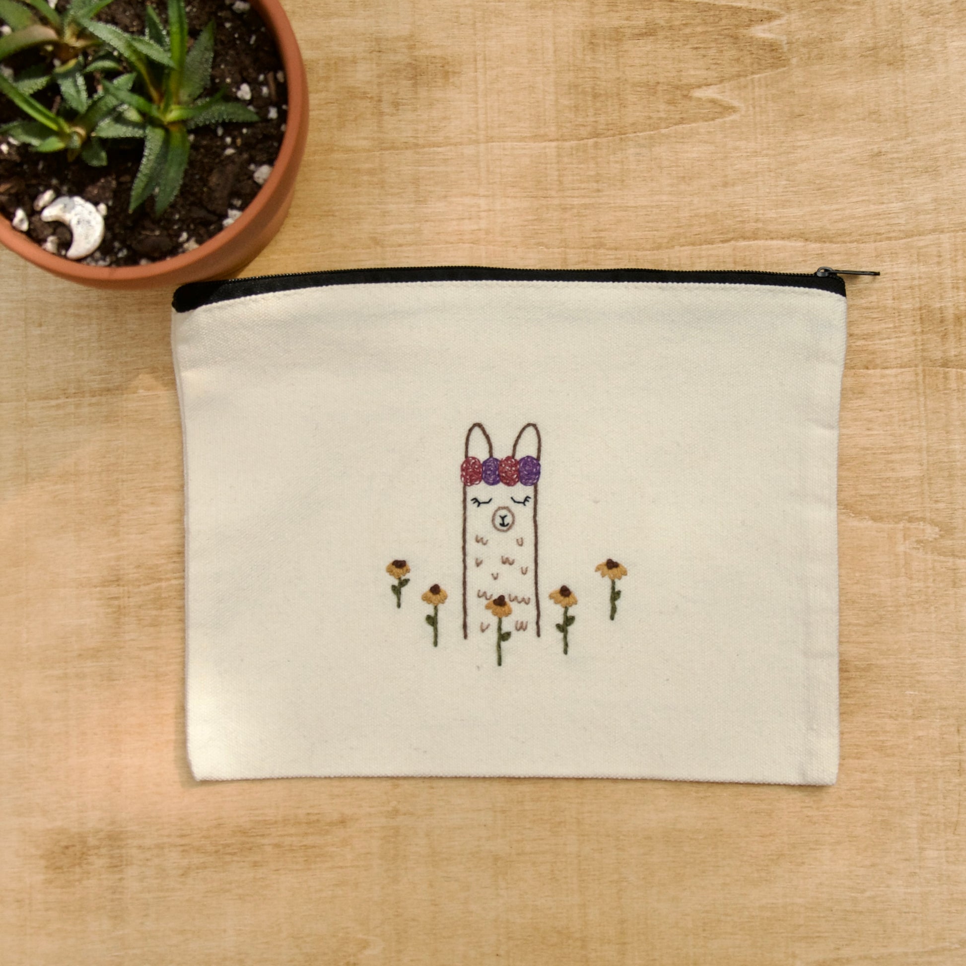 A white zip pouch with 1 embroidery alpaca with a flower crown  on a wooden surface with a potted plant nearby