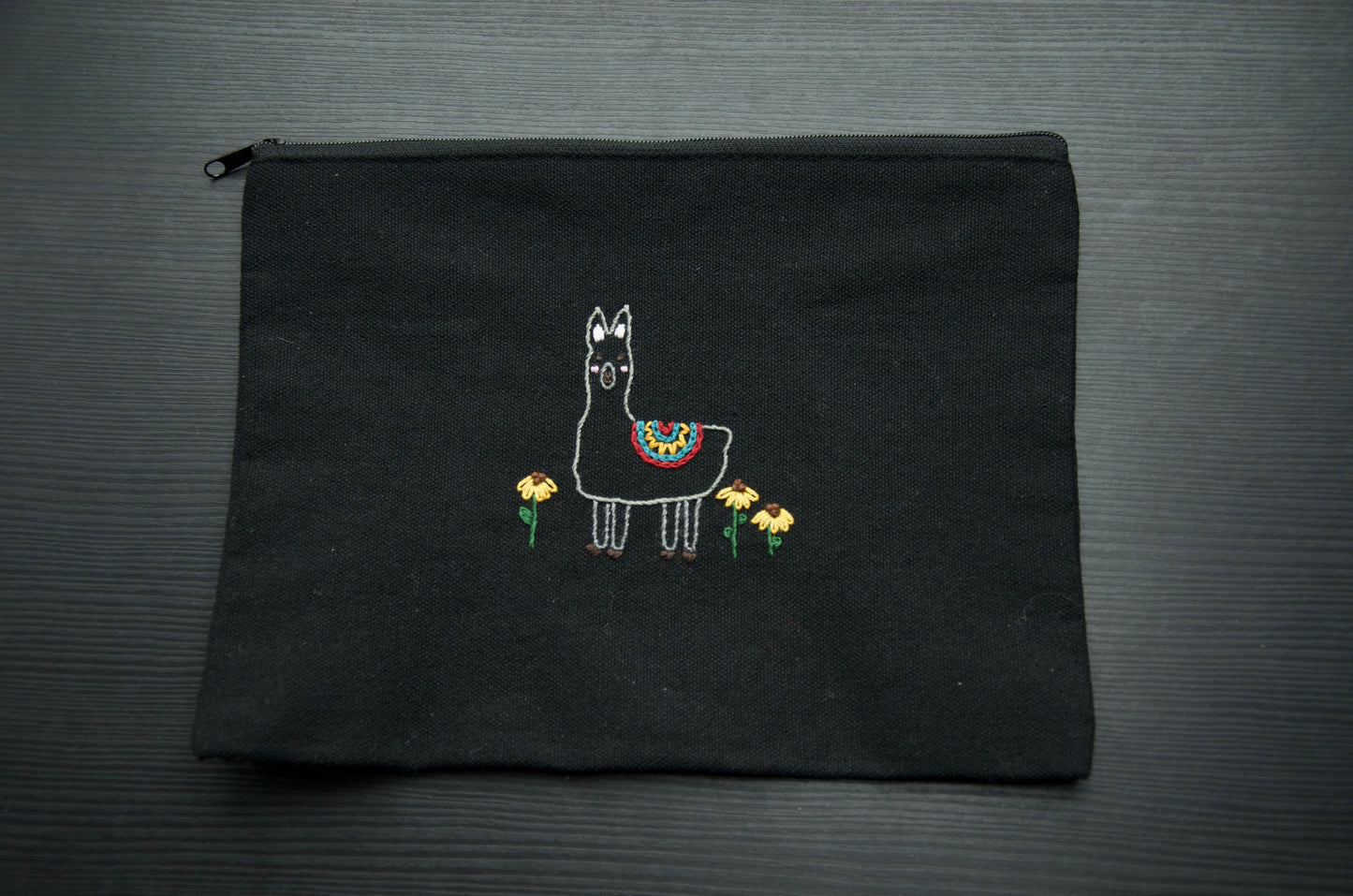 A black zip pouch with an embroidery alpaca in a flower field on a dark surface