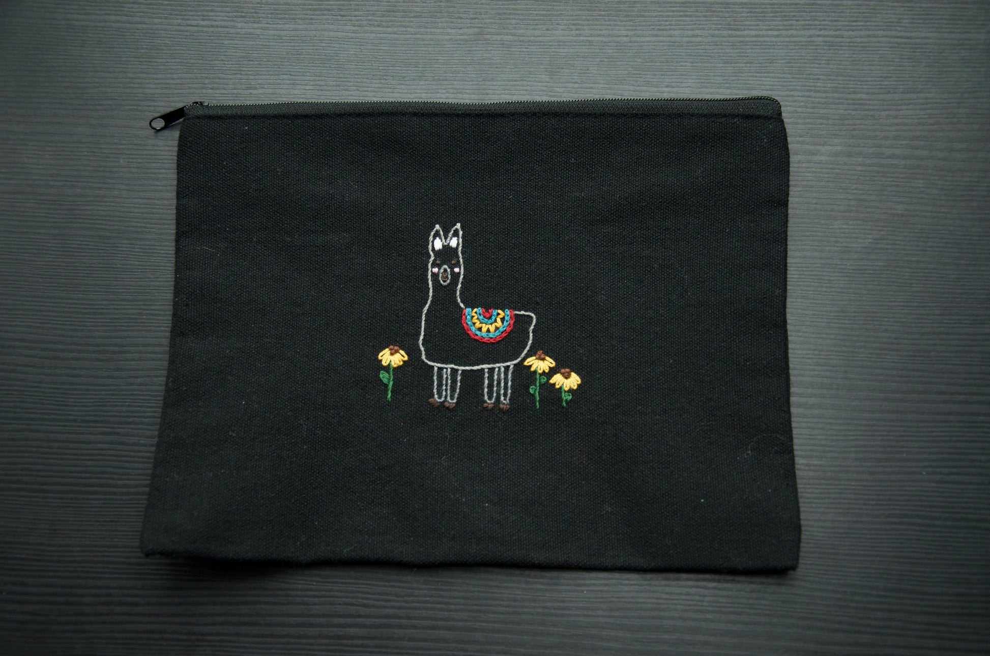 A black zip pouch with an embroidery alpaca in a flower field on a dark surface