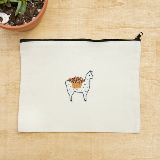 A white zip pouch with an embroidery alpaca with a flower basket on its' back  on a wooden surface with a potted plant nearby