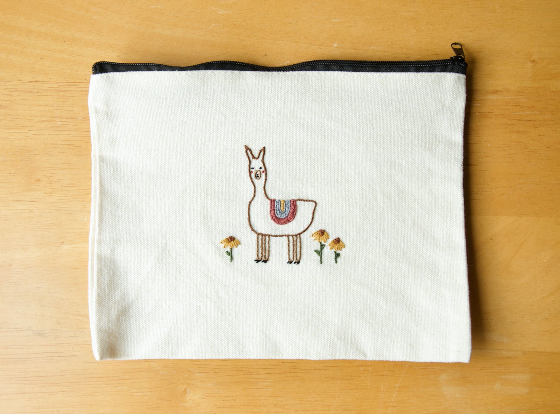 A white zip pouch with embroidery of an alpaca in a flower field on the front, placed on a wooden surface.
