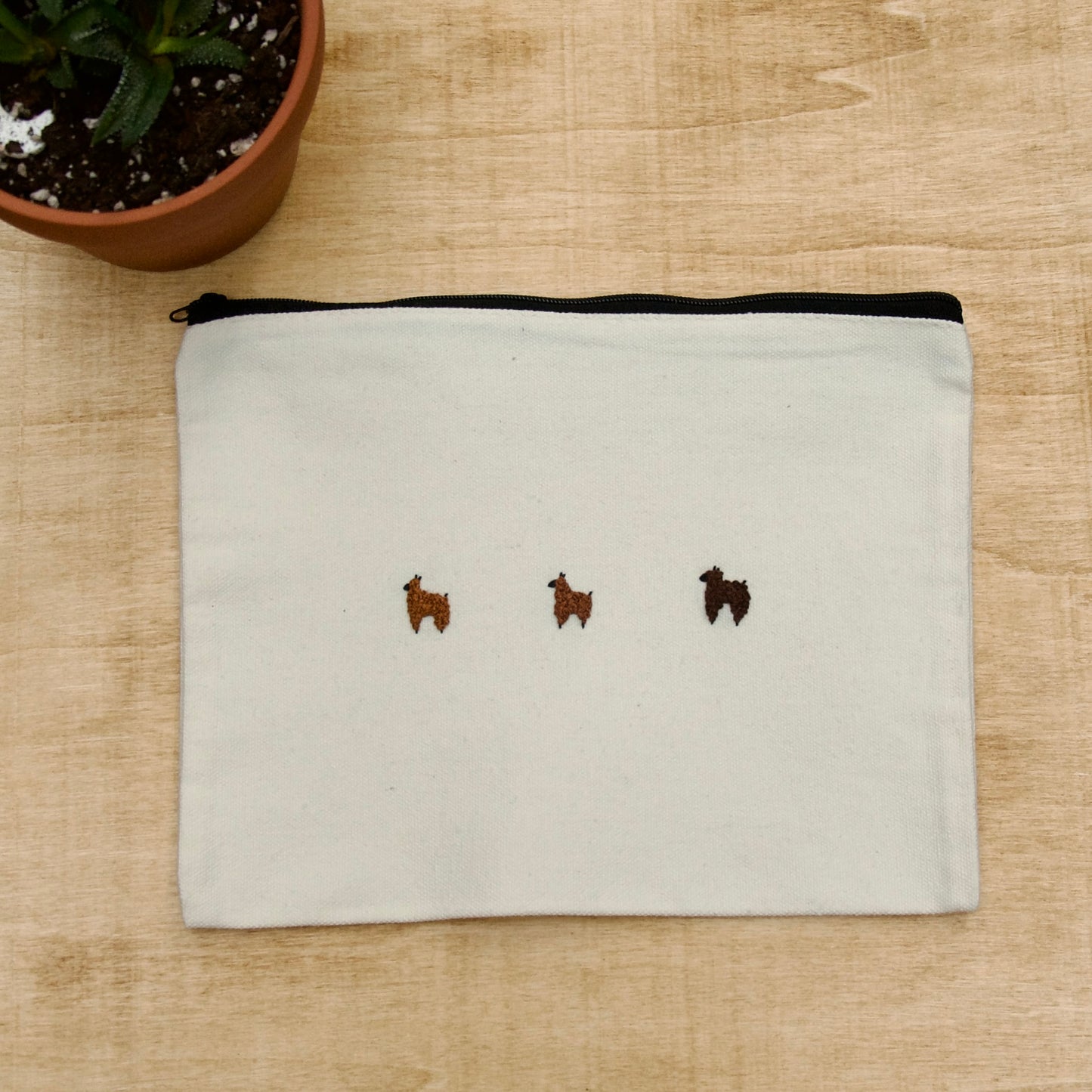 A white zip pouch with 3 brown embroidery alpacas on a wooden surface with a potted plant nearby