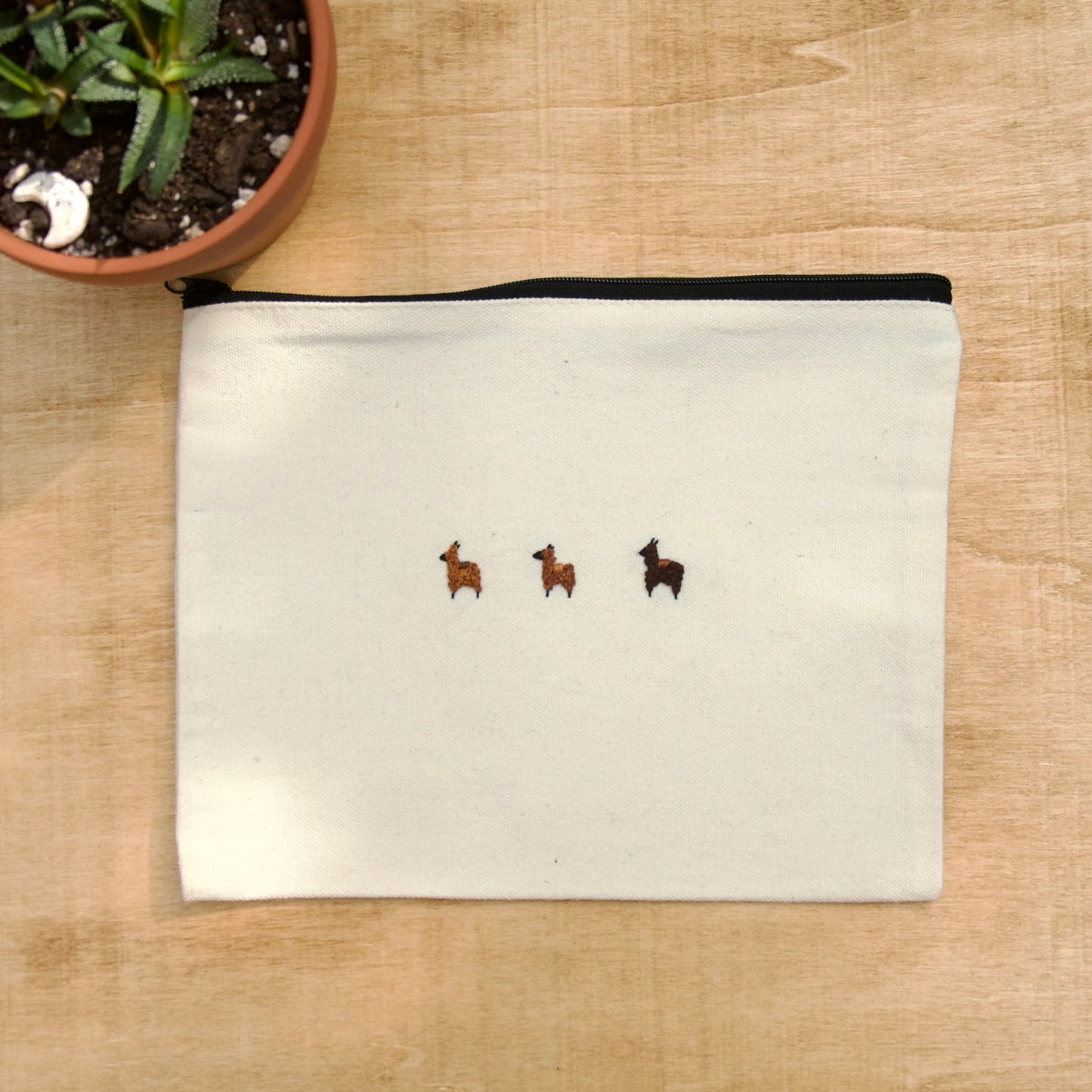 A white zip pouch with 3 brown embroidery alpacas on a wooden surface with a potted plant nearby