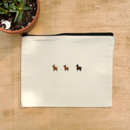 A white zip pouch with 3 brown embroidery alpacas on a wooden surface with a potted plant nearby