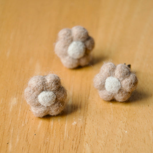 Three handmade felted air fresheners designed to clip to a car vent, shaped like flowers with a beige and white woollen texture, displayed on a wooden surface.