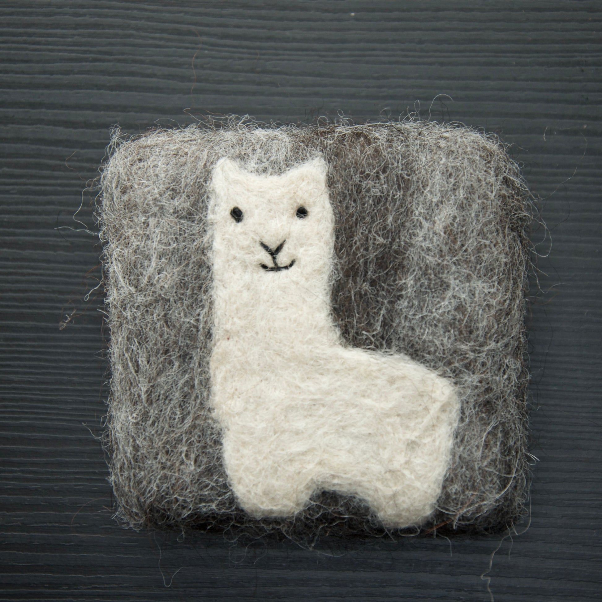 A felted soap bar shaped like an alpaca, enclosed in a square of brown felt, displayed on a dark background.