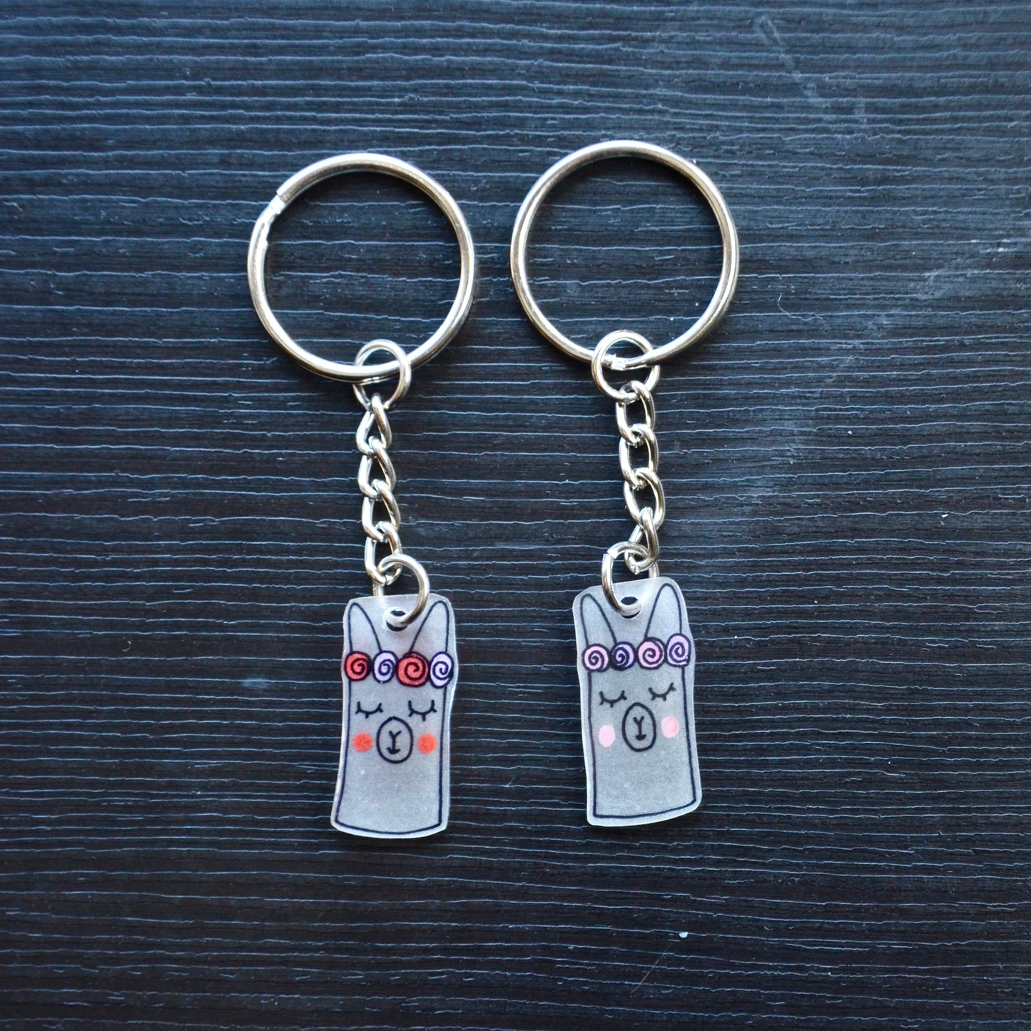 A collection of hand-drawn keychains with various cute designs, attached to silver split loops with chains. The keychains are made from recycled plastic and are in different colors, featuring simple, minimalistic alpaca designs.