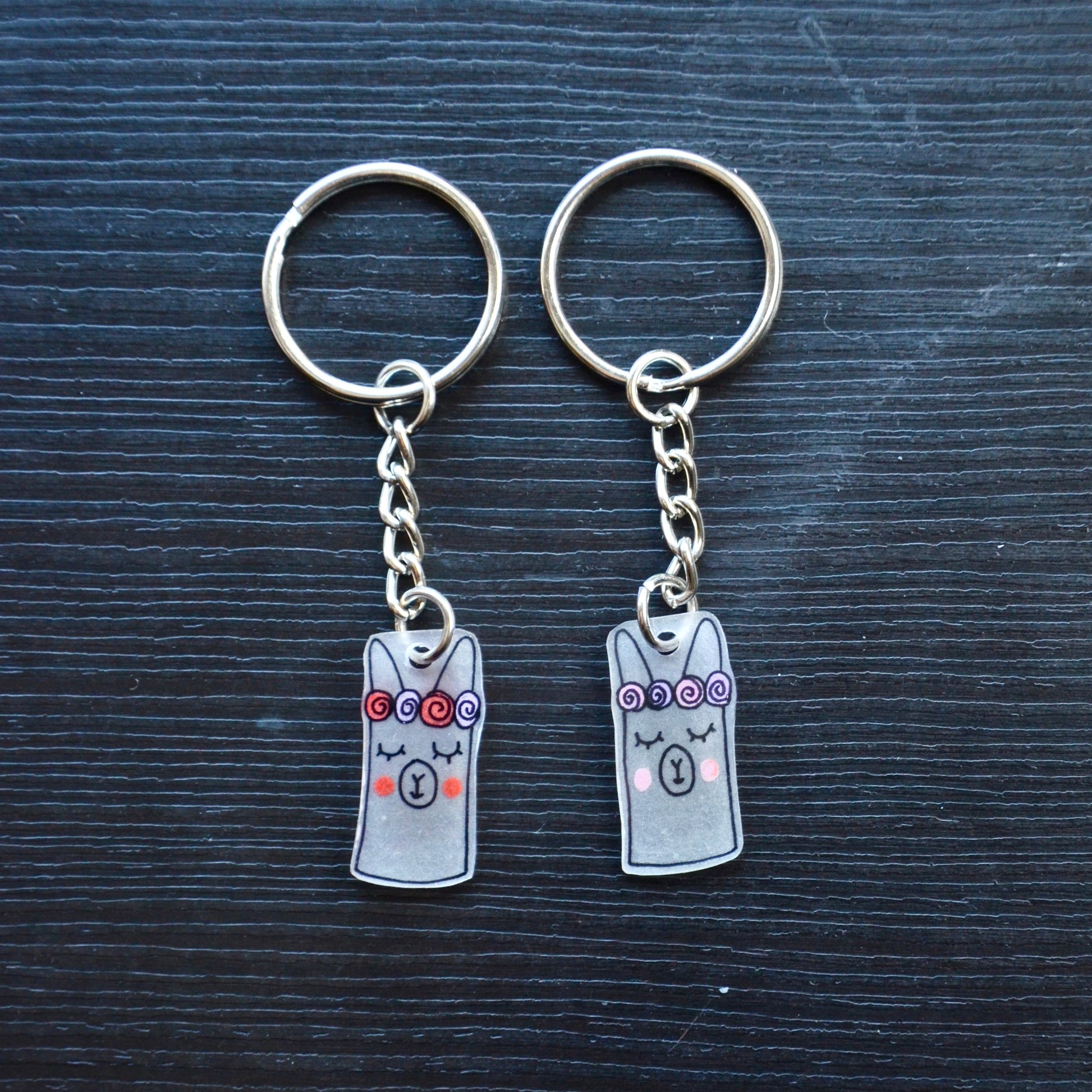 A collection of hand-drawn keychains with various cute designs, attached to silver split loops with chains. The keychains are made from recycled plastic and are in different colors, featuring simple, minimalistic alpaca designs.