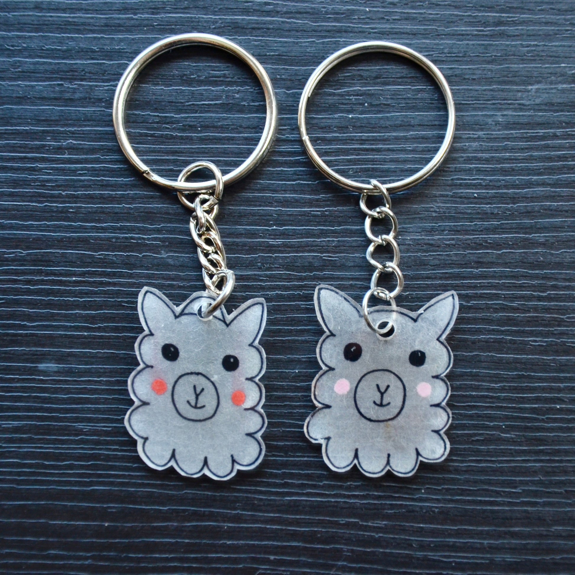 A collection of hand-drawn keychains with various cute designs, attached to silver split loops with chains. The keychains are made from recycled plastic and are in different colors, featuring simple, minimalistic alpaca designs.
