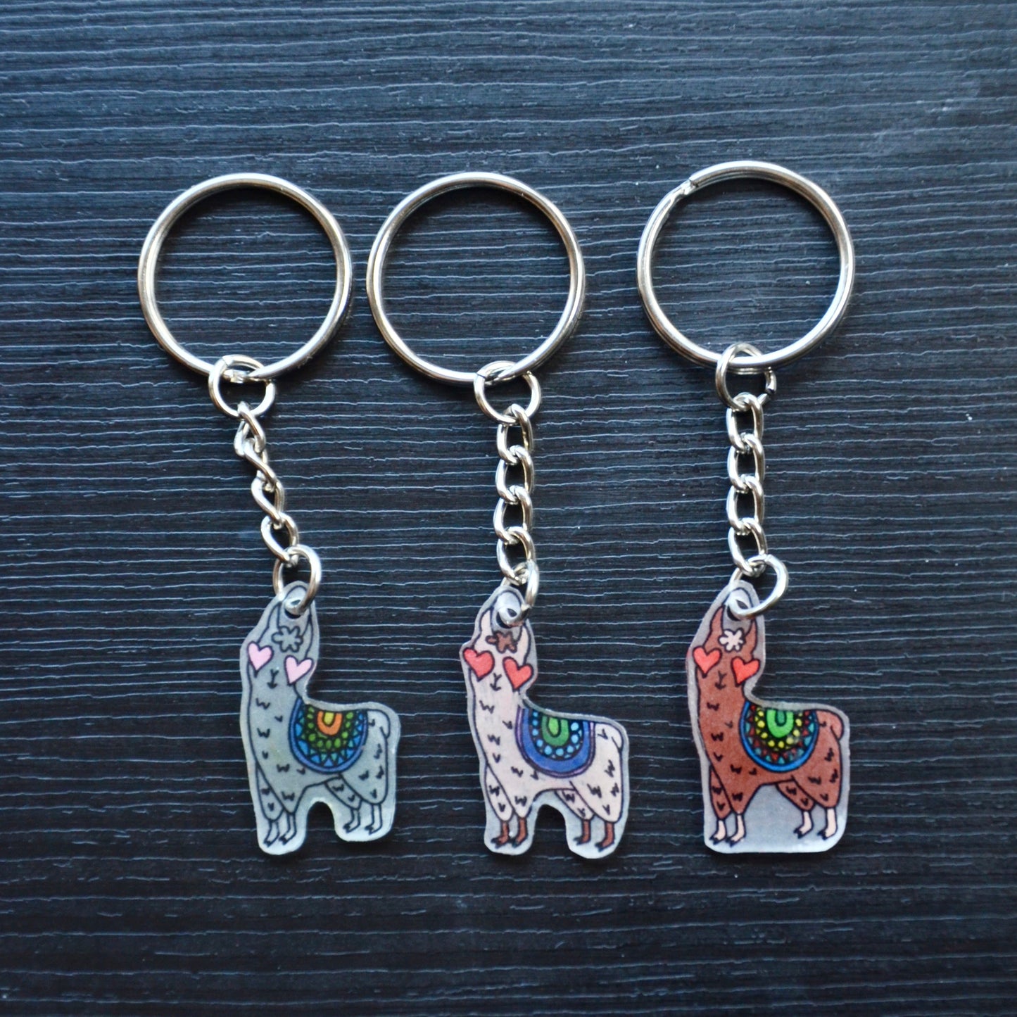 A collection of hand-drawn keychains with various cute designs, attached to silver split loops with chains. The keychains are made from recycled plastic and are in different colors, featuring simple, minimalistic alpaca designs.