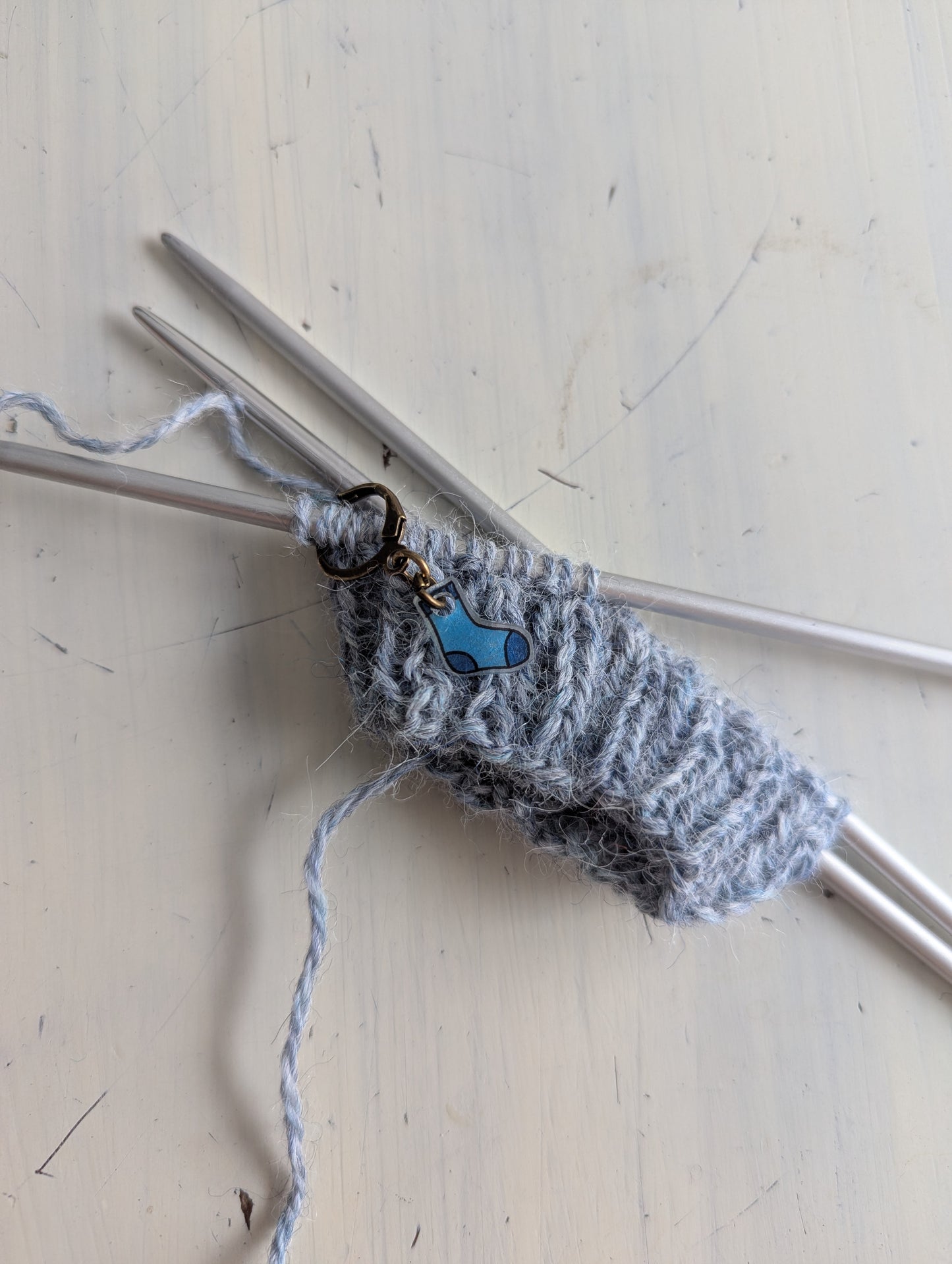 Knitted gray yarn on knitting needles with blue sock stitch minder on a light wooden surface