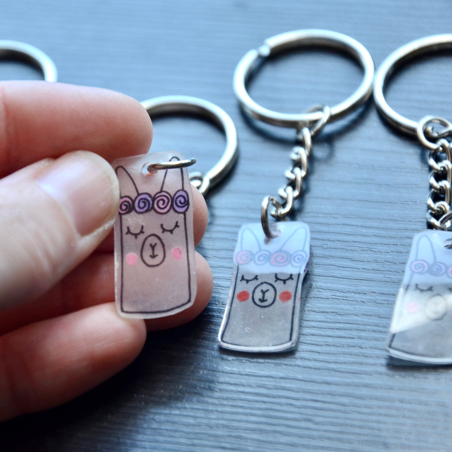 A collection of hand-drawn keychains with various cute designs, attached to silver split loops with chains. The keychains are made from recycled plastic and are in different colors, featuring simple, minimalistic alpaca designs.