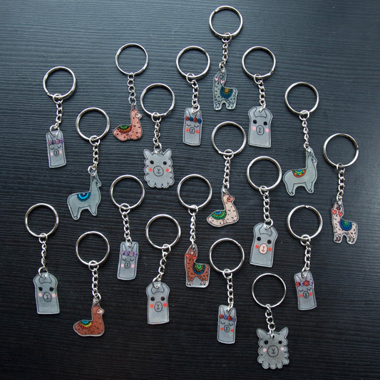 A collection of hand-drawn keychains with various cute designs, attached to silver split loops with chains. The keychains are made from recycled plastic and are in different colors, featuring simple, minimalistic alpaca designs.