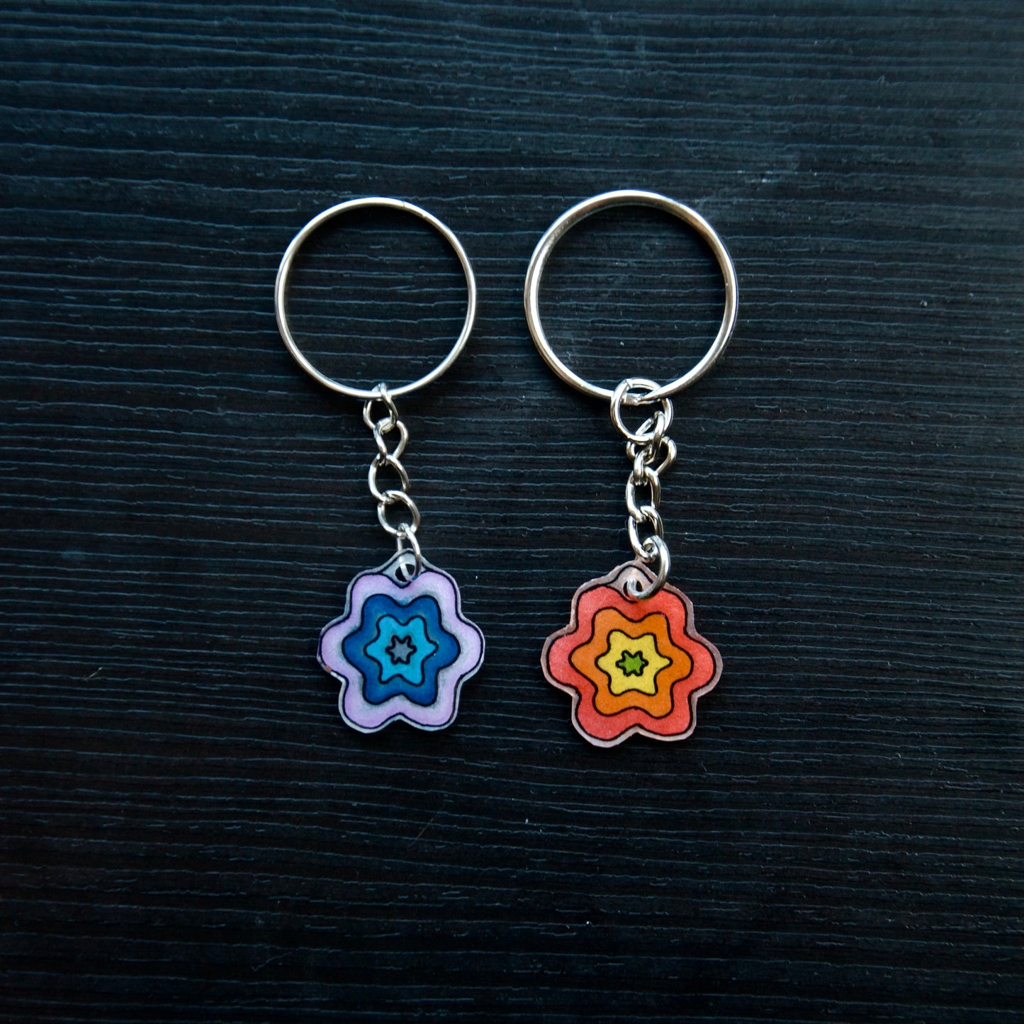 Two keychains with flower designs, one with a blue-pink gradient and the other with a yellow-orange gradient, attached to silver split loops with chains on a dark background.