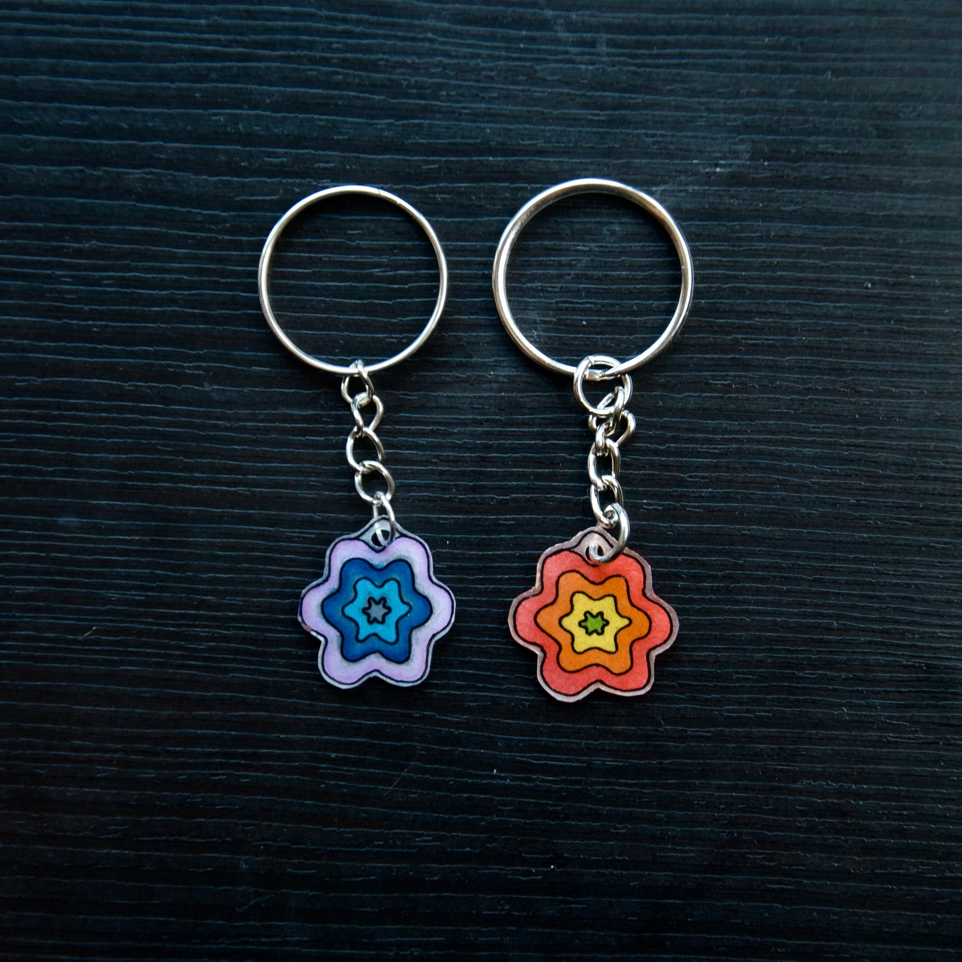 Two keychains with flower designs, one with a blue-pink gradient and the other with a yellow-orange gradient, attached to silver split loops with chains on a dark background.