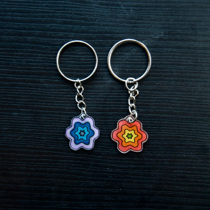 Two keychains with flower designs, one with a blue-pink gradient and the other with a yellow-orange gradient, attached to silver split loops with chains on a dark background.