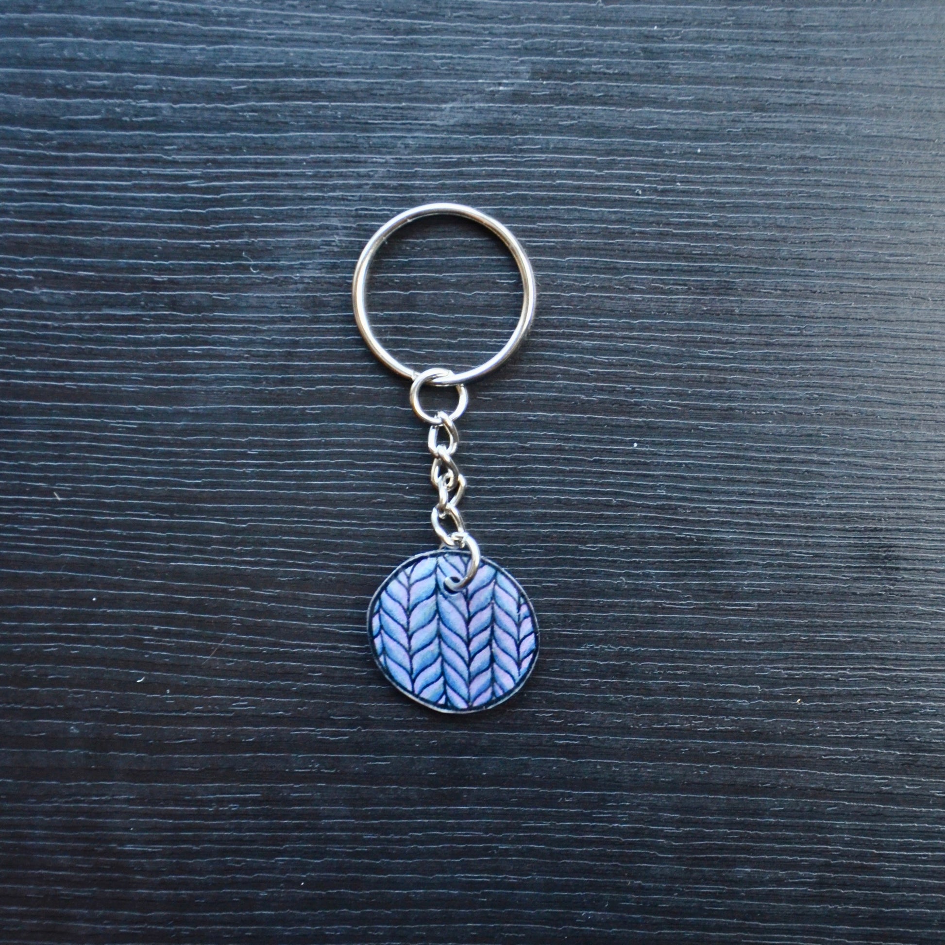 A set of colorful hand-drawn keychains made from recycled plastic, displayed on a dark wooden surface. Each keychain has a silver split loop and chain attachment.
